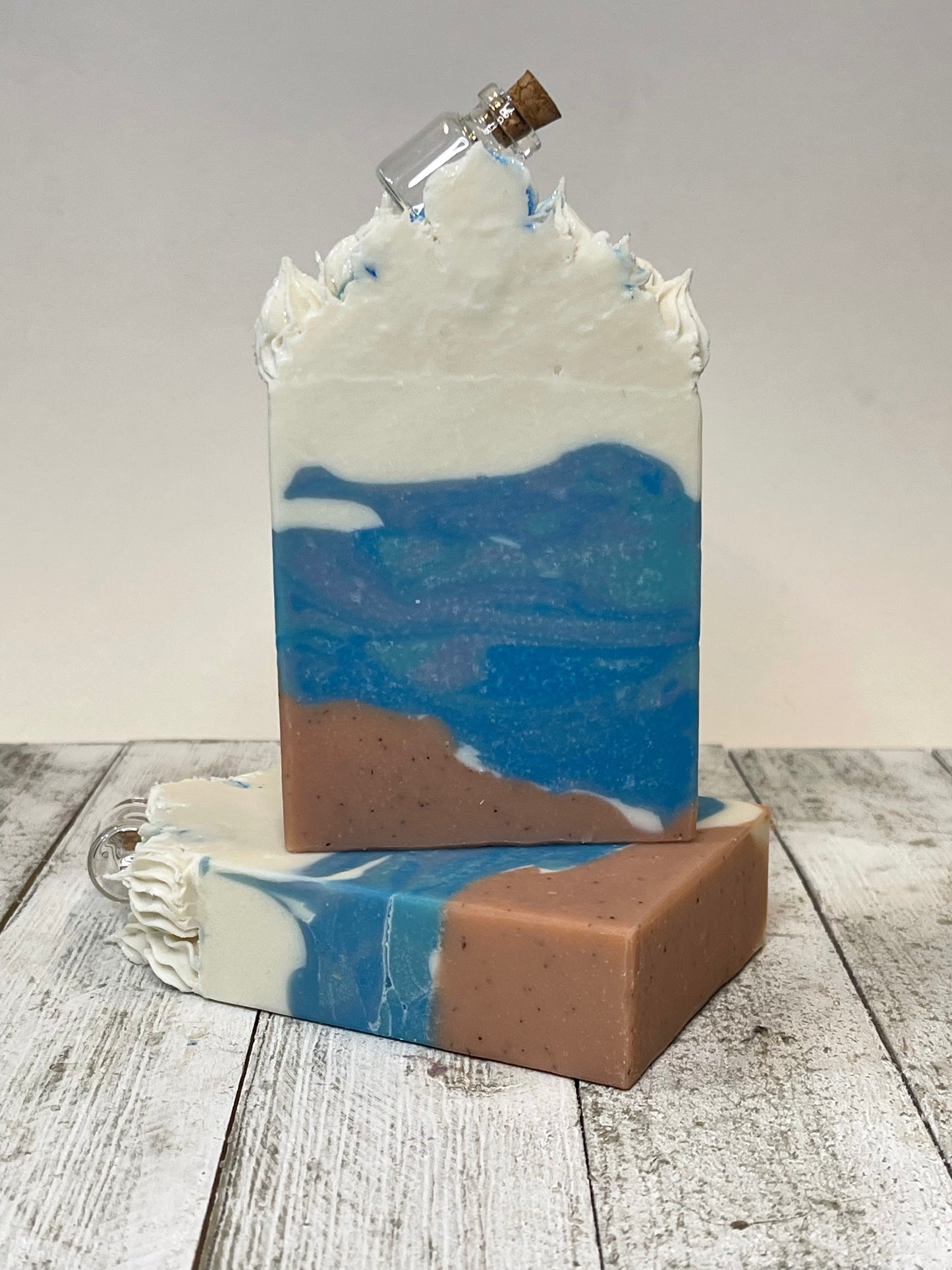 Message in the Bottle Soap Bar looks like a beach theme. There is brownish on the bottom that symbolizes the sand and different hues of blue for the ocean. The stark white symbolizes the waves crest. On top of the piping is the glass bottle with a cork.