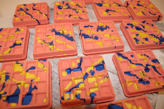 This picture has the waffle bath bombs in rows. They are a muted red color with blue and yellow color drizzled on top.